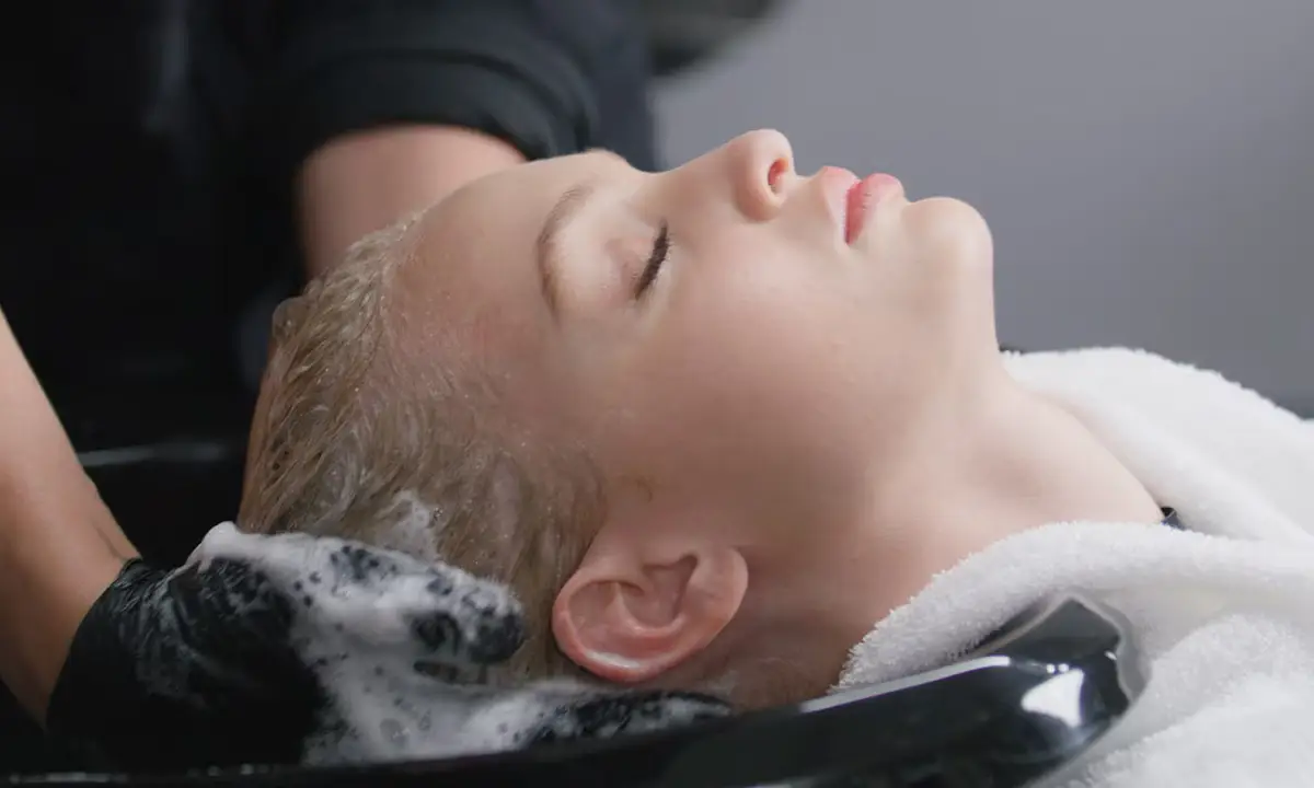 A person with closed eyes is having their hair washed at a salon sink with gloved hands applying shampoo and lather. | Anaala Salon and Spa, Madison WI