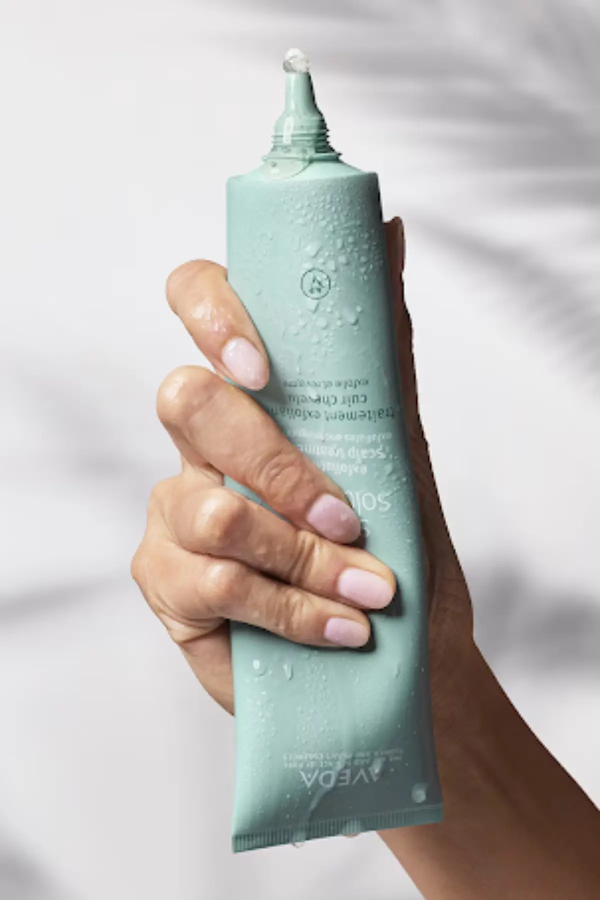 A hand with manicured nails holds a wet, mint-green tube of Aveda cream with a small drop of product at the tip of the open nozzle. | Anaala Salon and Spa, Madison WI