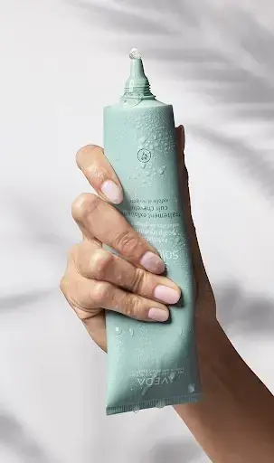 A hand holding a light blue tube of skincare product with a small amount squeezed out of the nozzle—perfect for those who believe Aveda rituals don't just elevate beauty, they create moments of mindfulness and renewal. | Anaala Salon and Spa, Madison WI