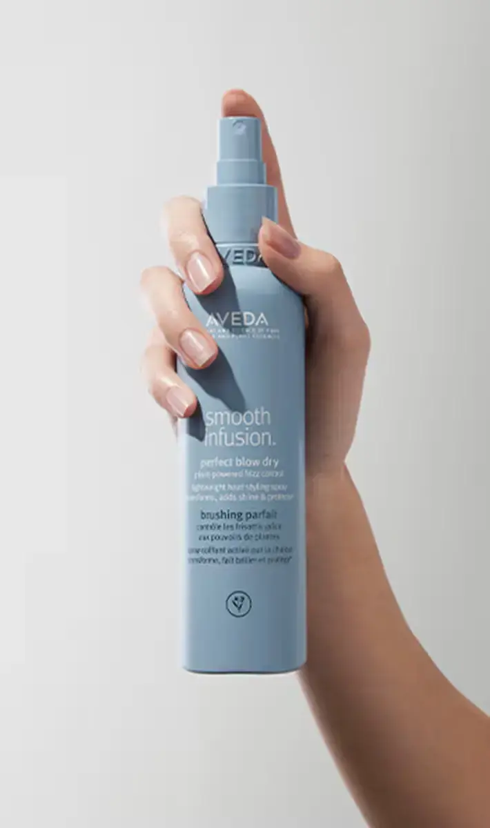 A hand holds a blue bottle of Aveda Smooth Infusion Perfect Blow Dry spray against a plain light background, reflecting how Aveda rituals don't just elevate beauty—they create moments of mindfulness and renewal. | Anaala Salon and Spa, Madison WI