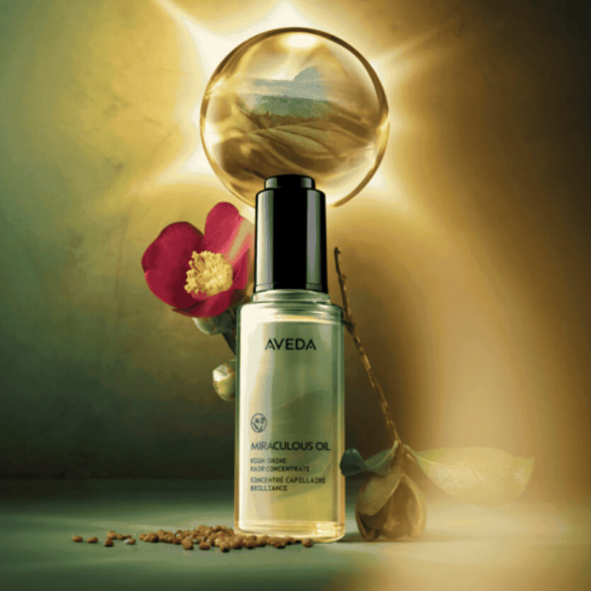 A bottle of Aveda Miraculous Oil is displayed in front of a glowing sphere, with a pink flower and seed pods beside it. | Anaala Salon and Spa, Madison WI
