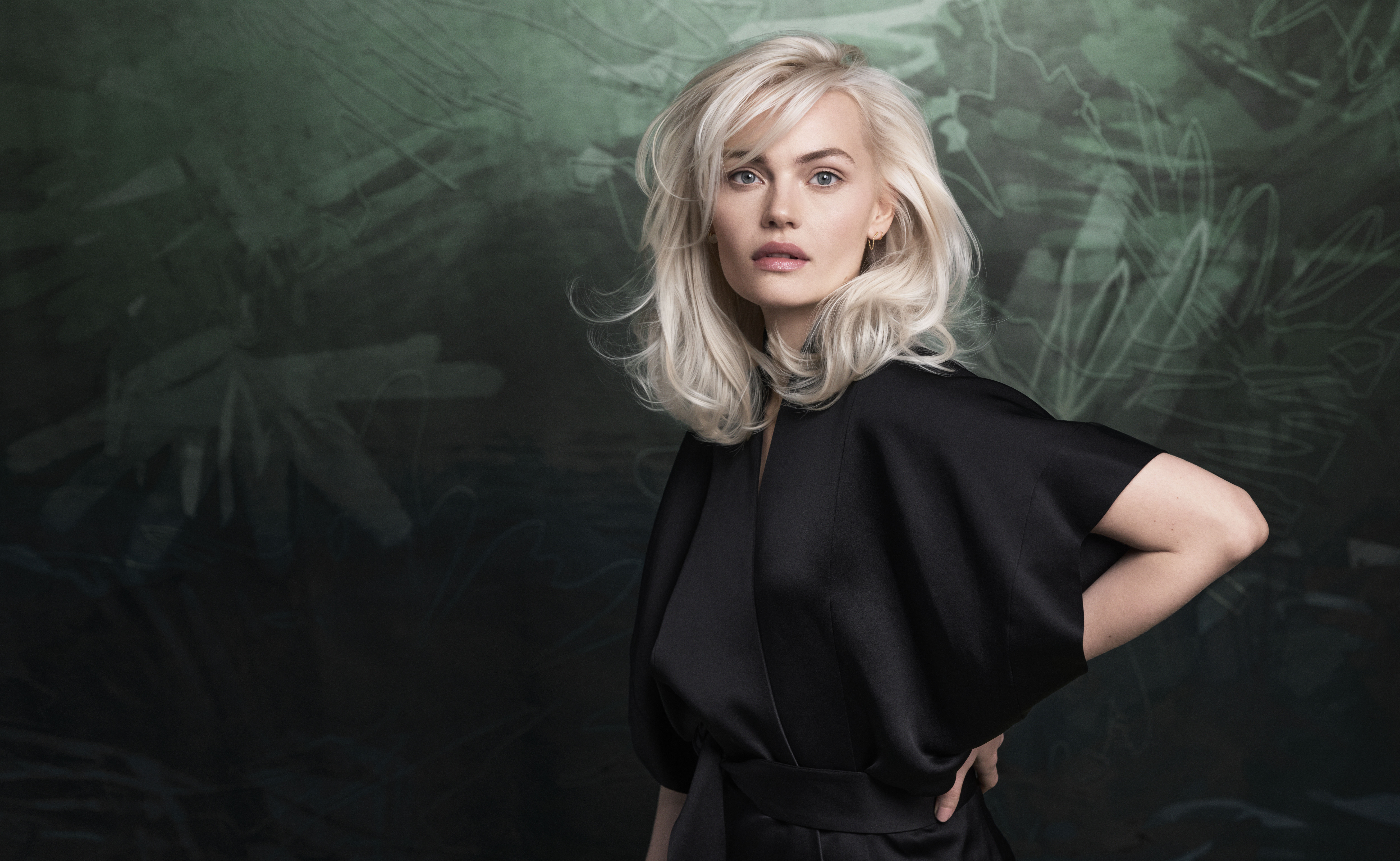 A woman with platinum blonde hair wearing a black outfit stands in front of a dark green, abstract background, looking at the camera with a neutral expression. | Anaala Salon and Spa, Madison WI