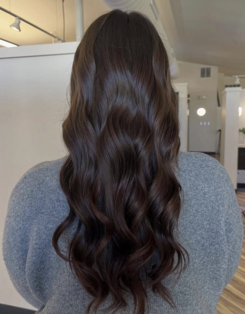 Person with long, wavy brown hair wearing a gray sweater, shown from the back in an indoor setting—ideal inspiration for hair color specialists Madison WI. | Anaala Salon and Spa, Madison WI