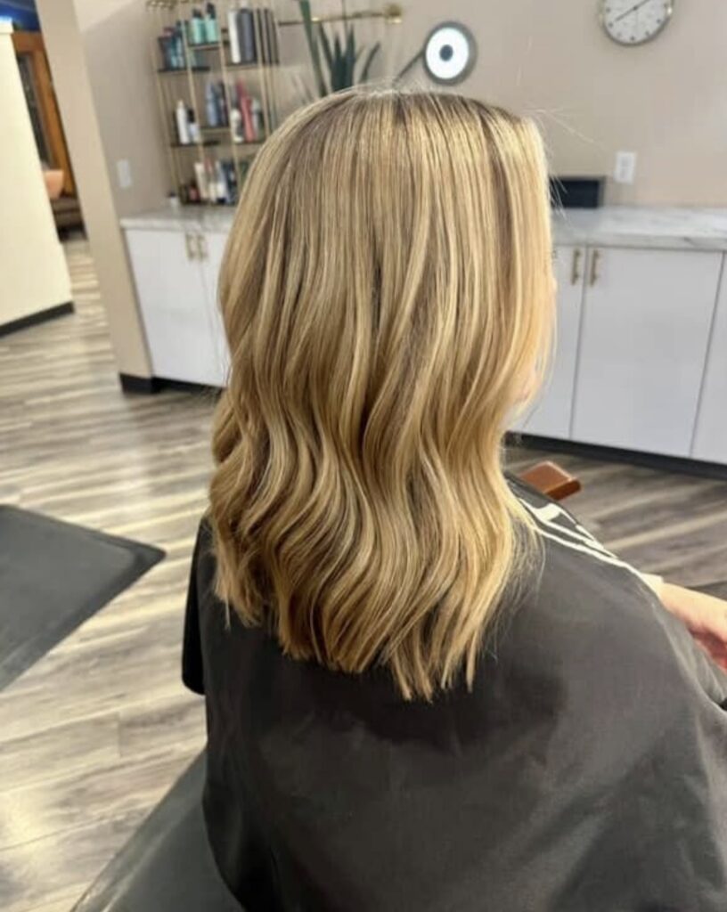 A person with shoulder-length, wavy blonde hair sits in a salon chair, facing away from the camera at one of the top hair color specialists Madison WI. Shelves with hair products and a clock are visible in the background. | Anaala Salon and Spa, Madison WI