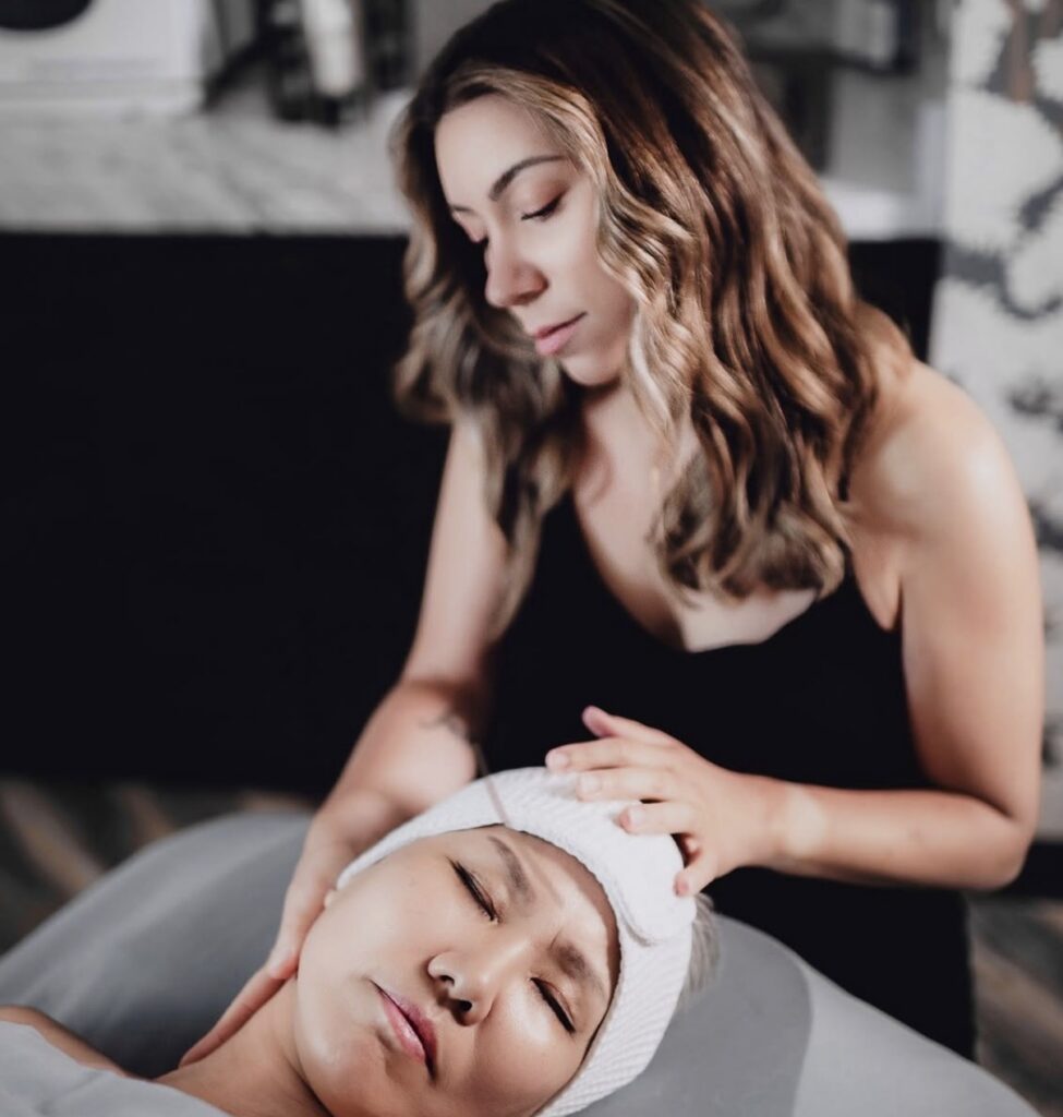 A woman with wavy hair, known for scalp health treatment Madison WI, gives a facial massage to another woman lying down with her eyes closed and a headband on. | Anaala Salon and Spa, Madison WI