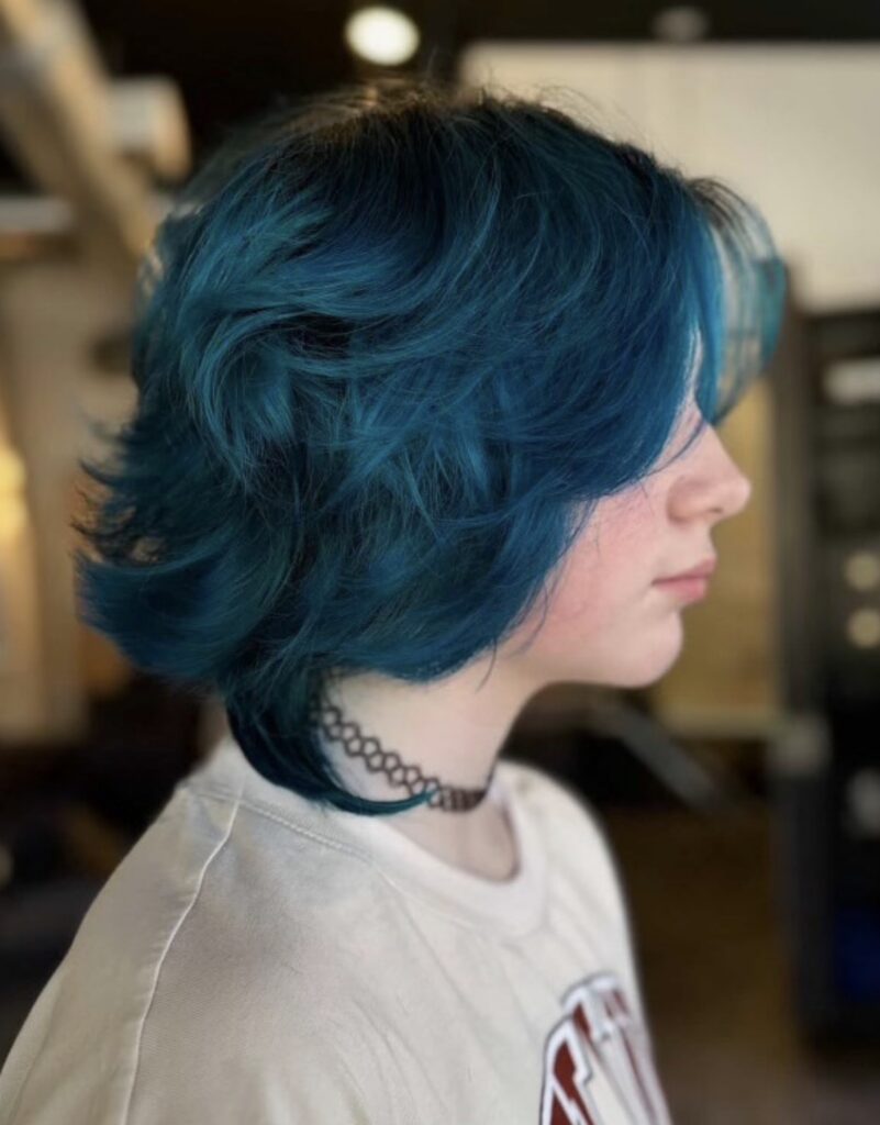 Person with layered, chin-length teal hair—styled by hair color specialists in Madison WI—wearing a white t-shirt and a black choker, shown in profile against an indoor background. | Anaala Salon and Spa, Madison WI