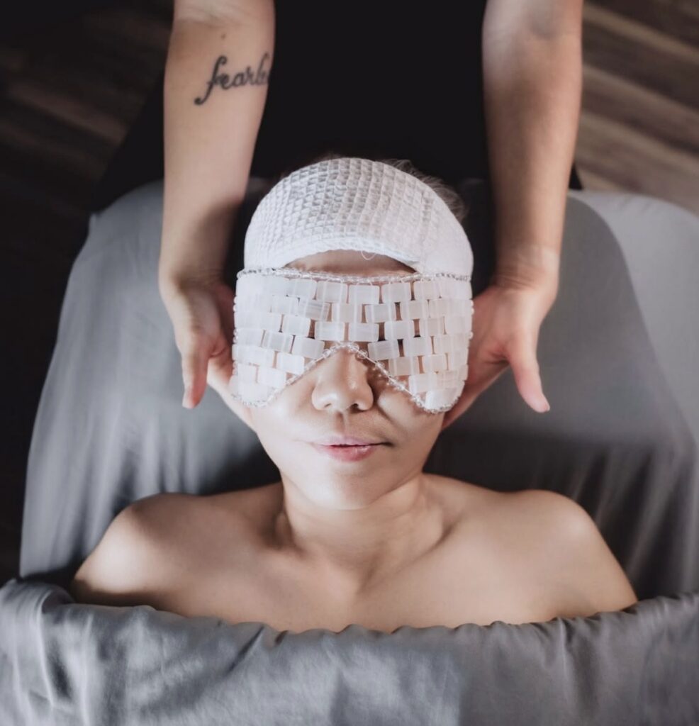 A person lies under a gray blanket with a woven eye mask and head wrap, while another's hands gently cradle their head—an experience of scalp health treatment Madison WI guests love. | Anaala Salon and Spa, Madison WI