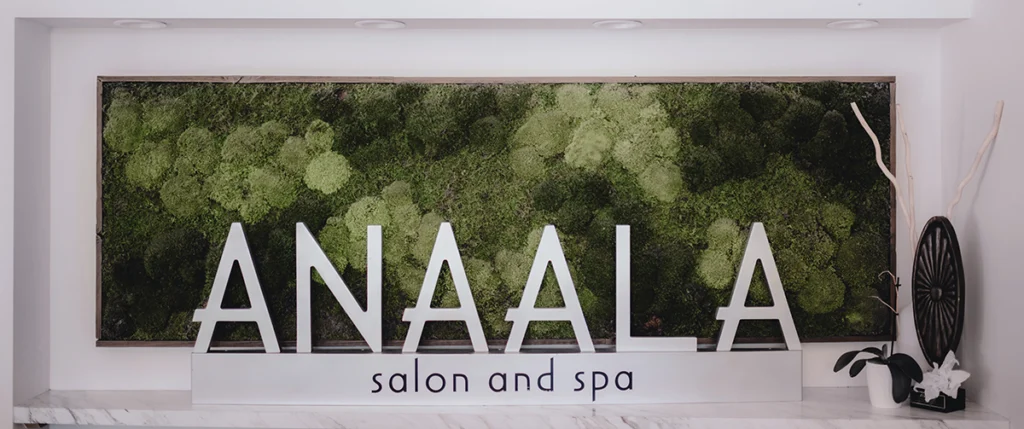 A sign reading "ANAALA salon and spa" is displayed in front of a green moss wall with decorative branches and a black circular ornament, reflecting the style of the best salon in Madison WI. | Anaala Salon and Spa, Madison WI