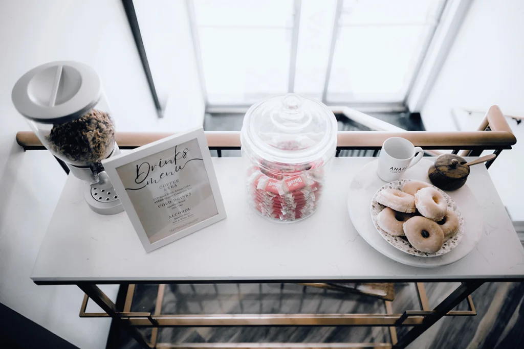 A countertop at the best salon in Madison WI features a cereal dispenser, framed drink menu, glass jar of pink snacks, plate of donuts, mug, and a small creamer pitcher for guests to enjoy. | Anaala Salon and Spa, Madison WI