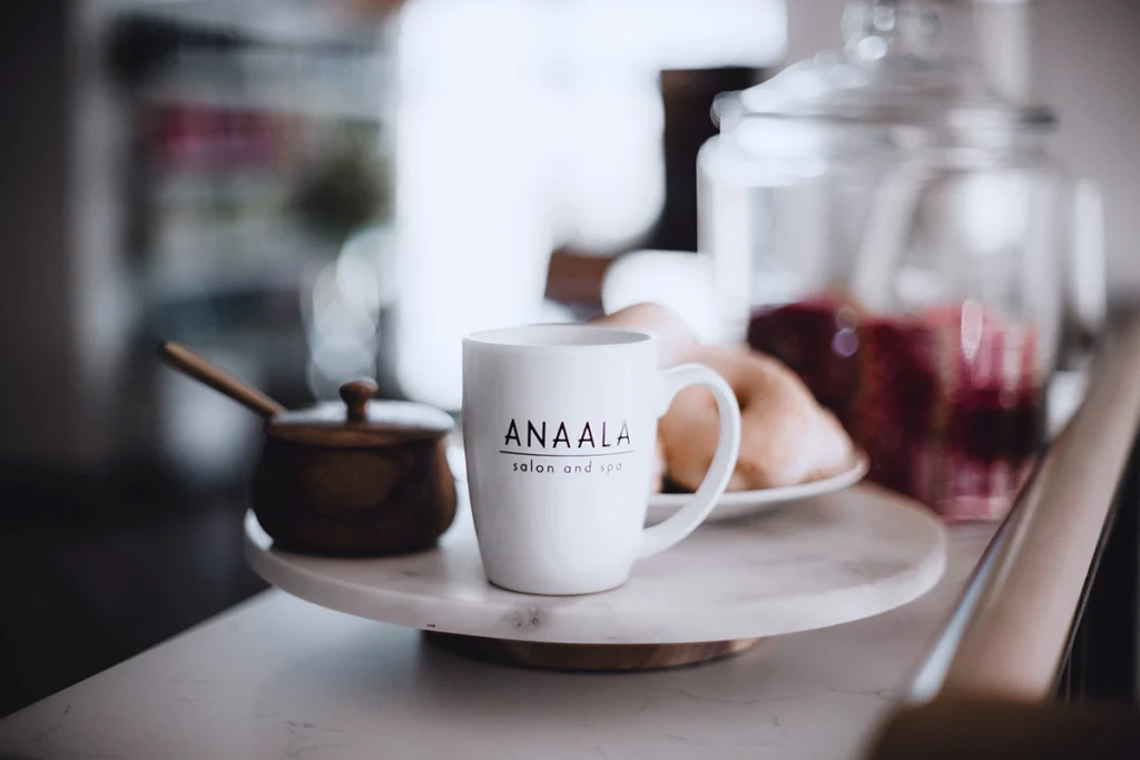 A white mug with "ANAALA salon and spa" printed on it sits on a plate next to a wooden container and pastries—perfect for relaxing after visiting the hair color specialists Madison WI trusts. | Anaala Salon and Spa, Madison WI