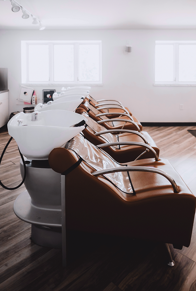 Row of empty salon shampoo chairs and sinks in a modern, well-lit room with wooden flooring—your destination for scalp health treatment in Madison, WI—large windows provide a relaxing backdrop. | Anaala Salon and Spa, Madison WI